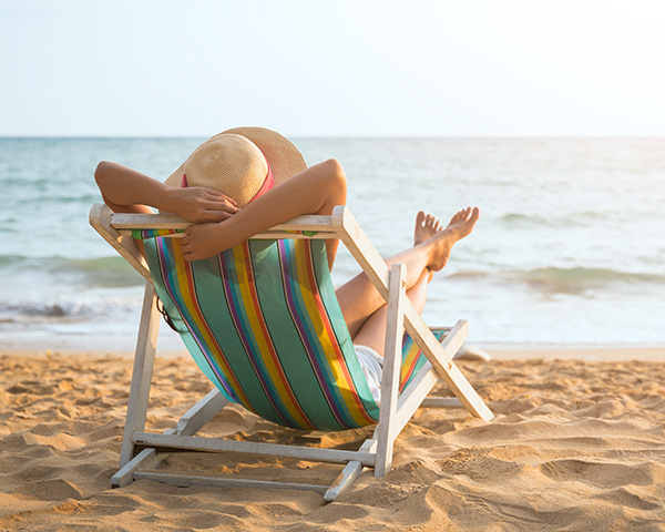 Person in a sun hat relaxing on a striped deck chair by the beach, facing the ocean.