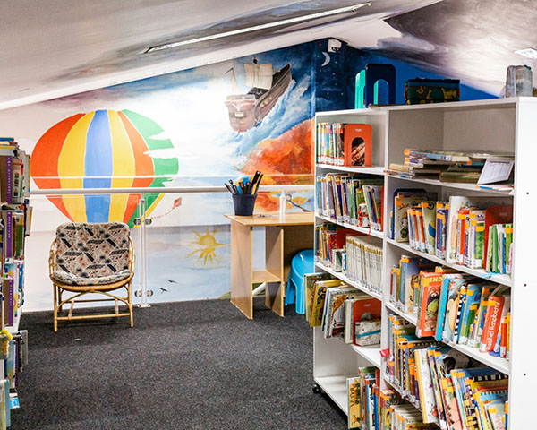 A cozy library corner with colorful bookshelves, a chair, and a mural of a hot air balloon and ship.