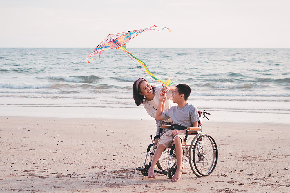 Two people, one in a wheelchair, are flying a colorful kite on a sandy beach near the ocean. They are smiling and enjoying the activity.