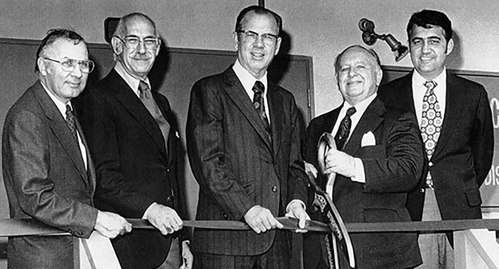 A group of five men in suits stand together at a ribbon-cutting ceremony. One man holds large scissors, all are smiling.