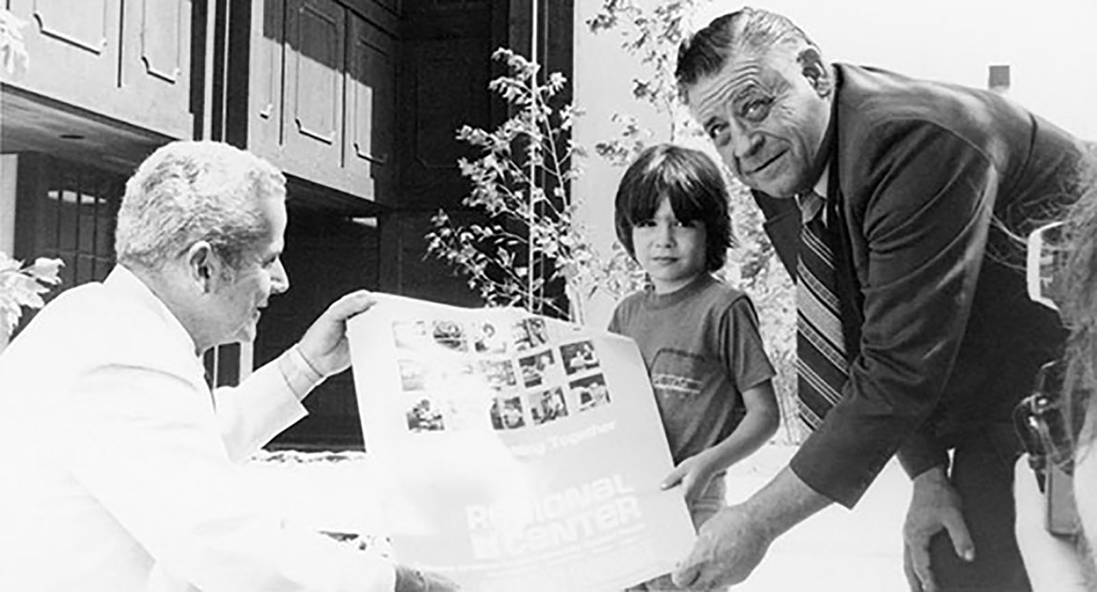 Two men and a child hold a large poster outdoors. One man is kneeling, and the other is standing beside the child.