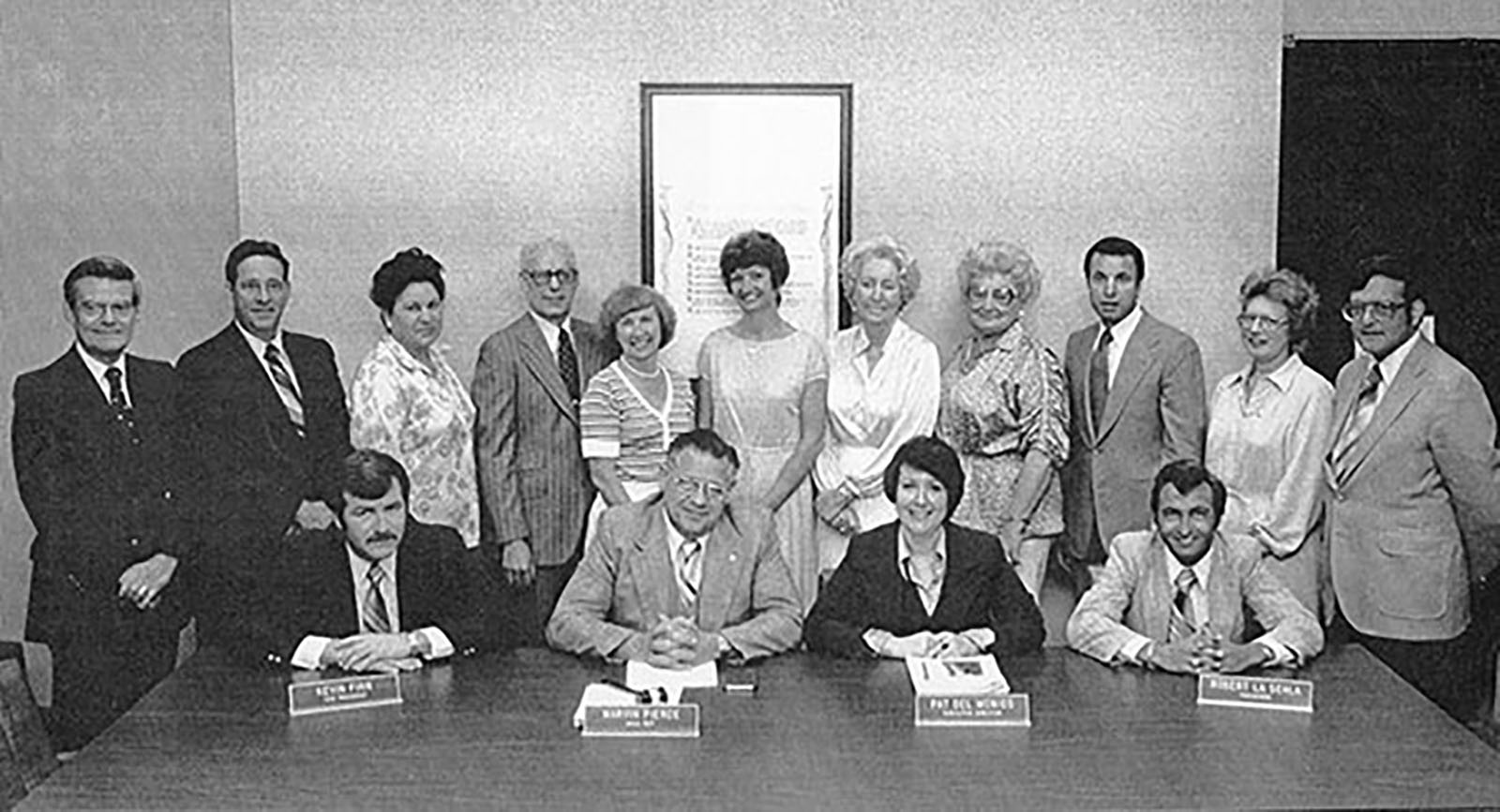 Group of fifteen people, including twelve standing and three seated at a table with nameplates, in a formal setting.