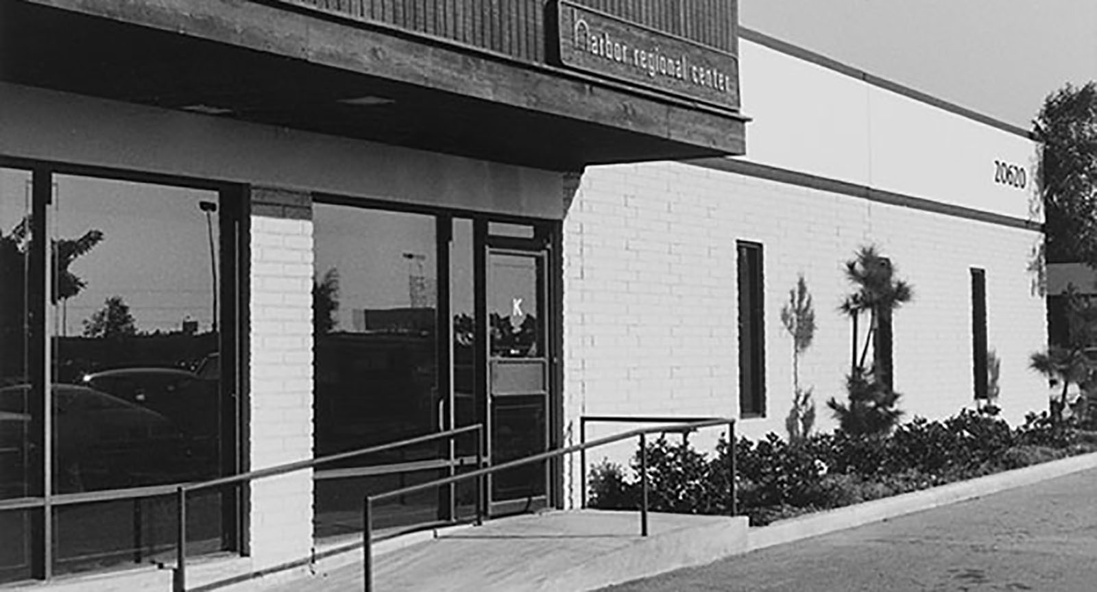 Black and white image of a commercial building with large windows, a ramp, and an entrance door. The sign above reads 