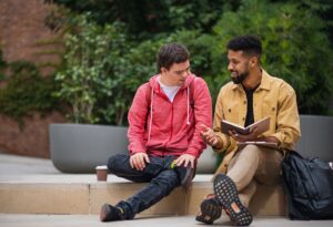 Two people sitting on a concrete ledge outside smiling and one is holding a notebook.