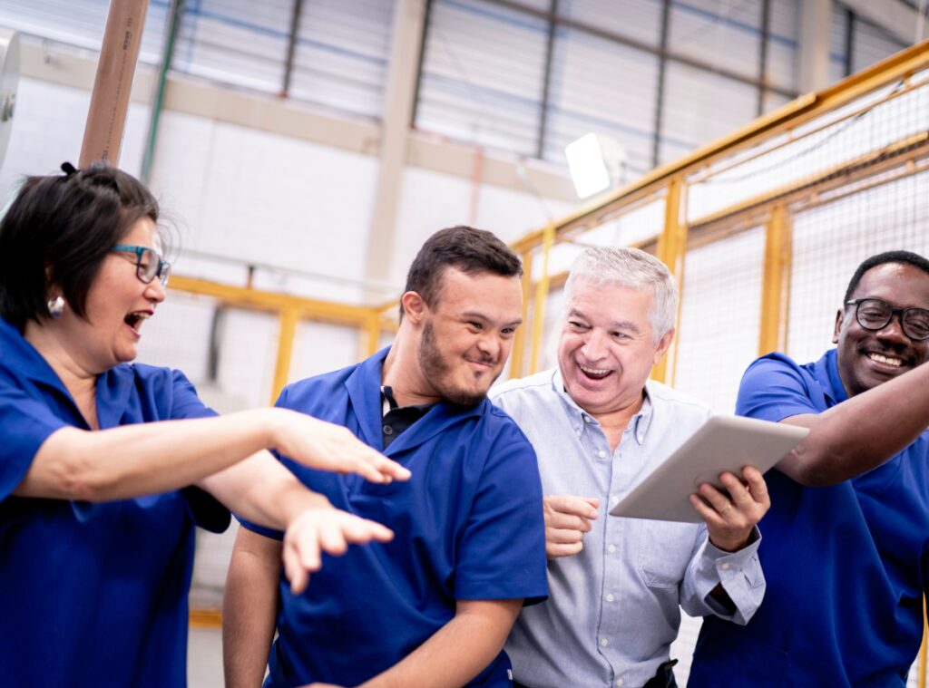 A group of four people laughing and smiling at each other. One person is holding a tablet. Wooden framed room is blurred in the background