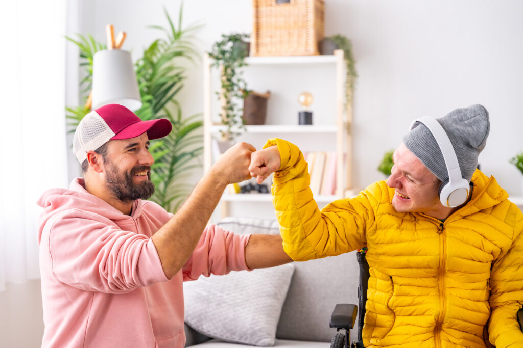 A man in a wheelchair wearing headphones and friend feeling happy and giving each other a fist bump while at home