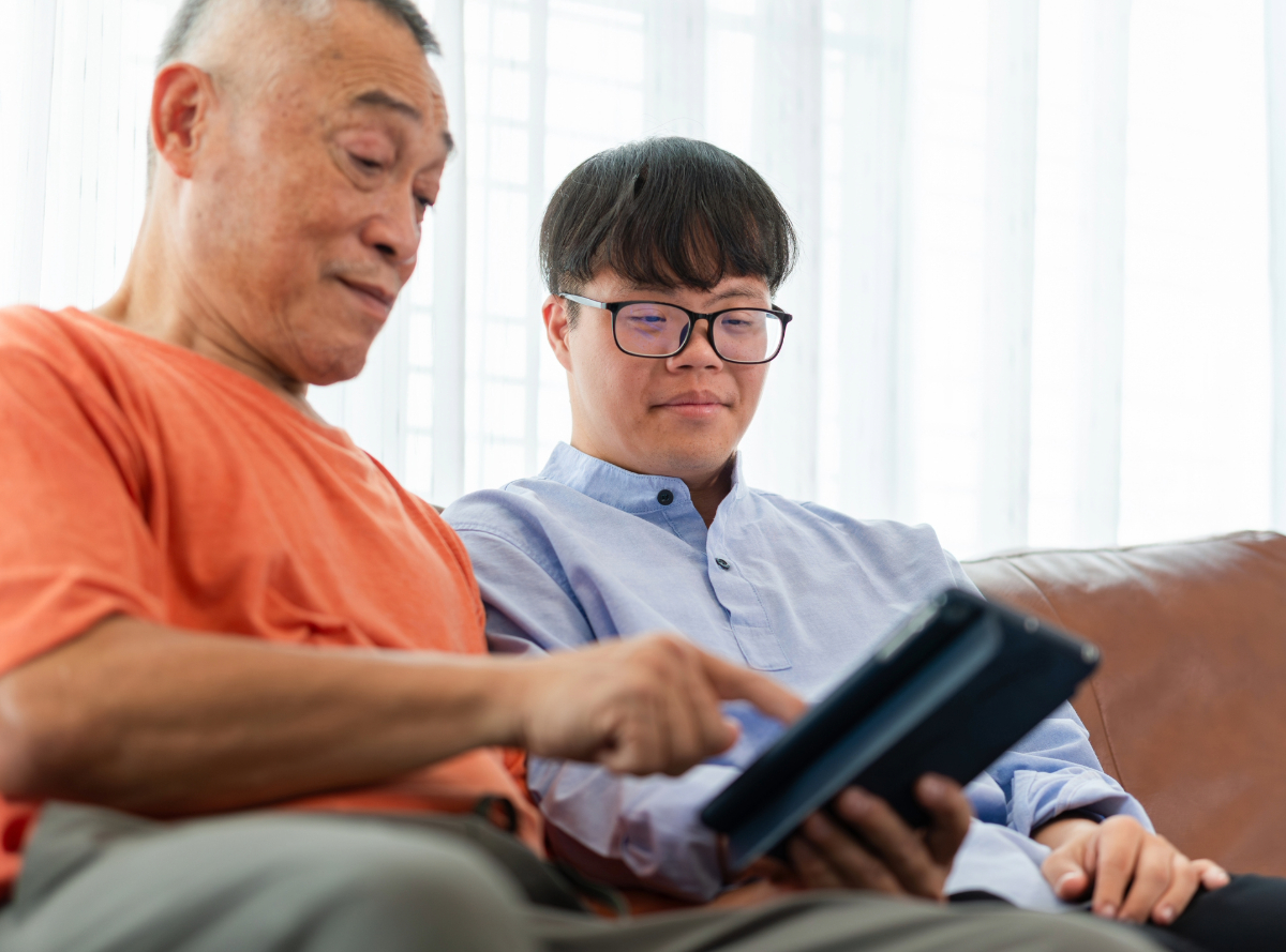 A young man with a developmental disability is sitting next to his father on a leather couch and they are looking at a tablet.