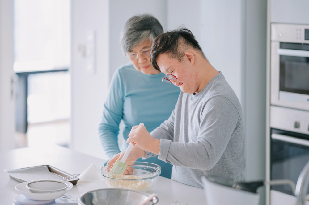 A young man wearing glasses is using a spatula to mix ingredients in a glass bowl while his grandmother is looking on. They are in the kitchen